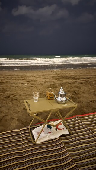 A beach with a table set up on the sand, featuring a teapot, a glass of water, and a striped blanket. 4