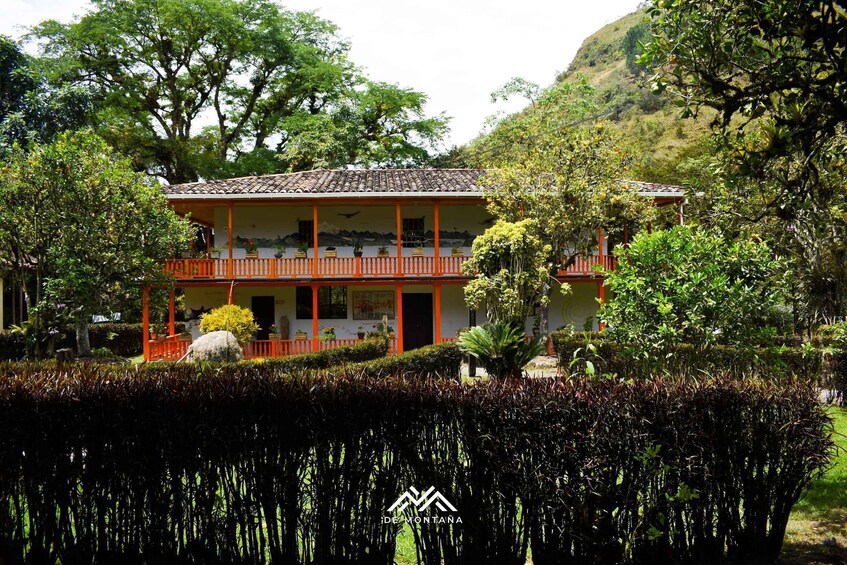 A two-story building with a red roof and orange railings, surrounded by lush greenery and trees. 2