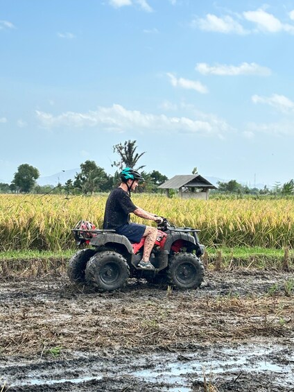 A person riding an ATV in a muddy field with a house and trees in the background. 3