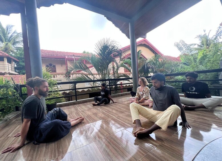 A group of people sitting on a tiled floor under a covered area with a view of a building with a red roof and palm trees. 1