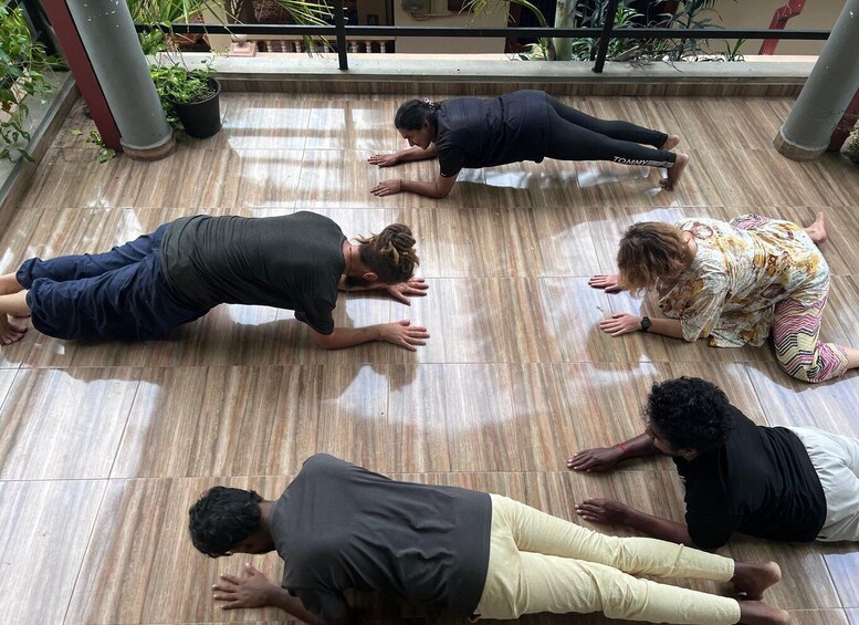 A group of people performing a floor exercise in an indoor setting with tiled flooring and potted plants in the background. 2