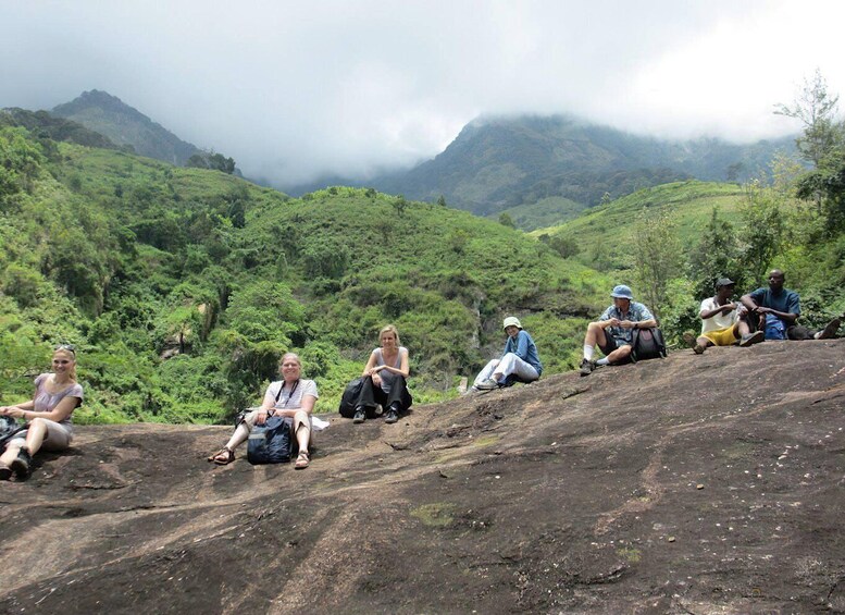 A group of people sitting on a rocky slope with lush green hills and mountains in the background. 1
