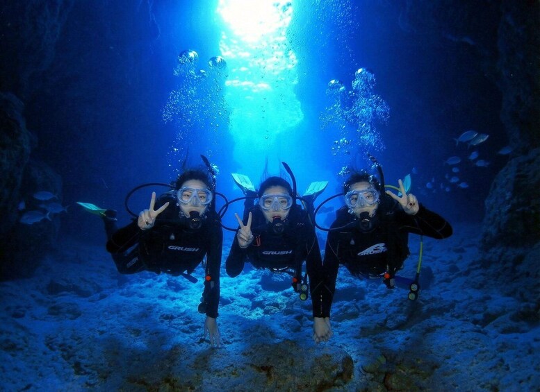 Three divers in black wetsuits with 'GRUSH' branding, making peace signs, underwater in a blue-lit environment with bubbles and fish. 5