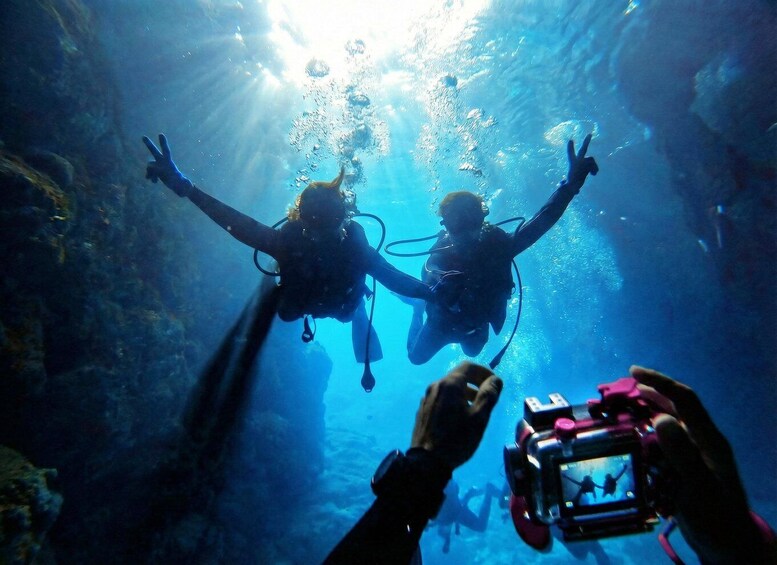 Two scuba divers underwater with their arms outstretched, surrounded by bubbles and sunlight filtering through the water. 1