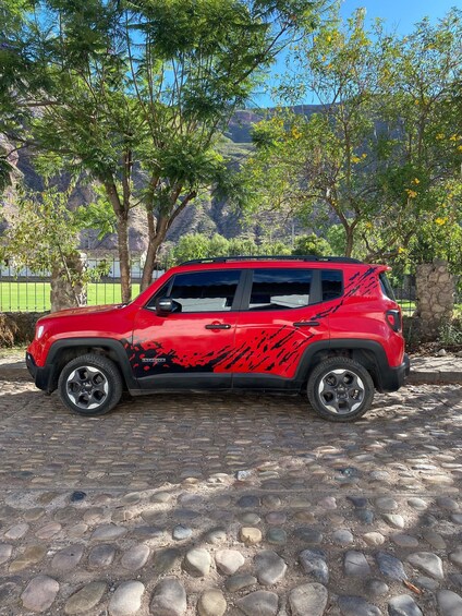 A red off-road vehicle parked on a cobblestone surface with trees and a mountainous background. 2