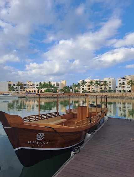 A wooden boat named 'HAWANA' docked at a pier with a backdrop of modern buildings and palm trees. 1