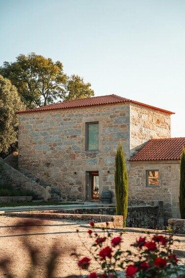 A stone building with a red-tiled roof, a window, and a door. There are trees and a clear sky in the background. 1