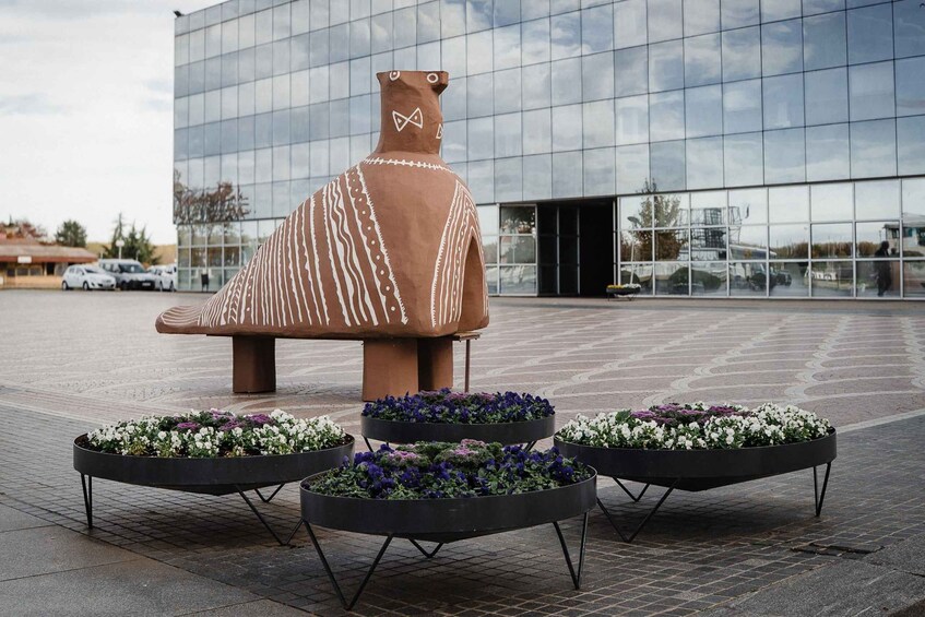 A modern sculpture of a seated figure with a patterned cloak, surrounded by flower planters. 4