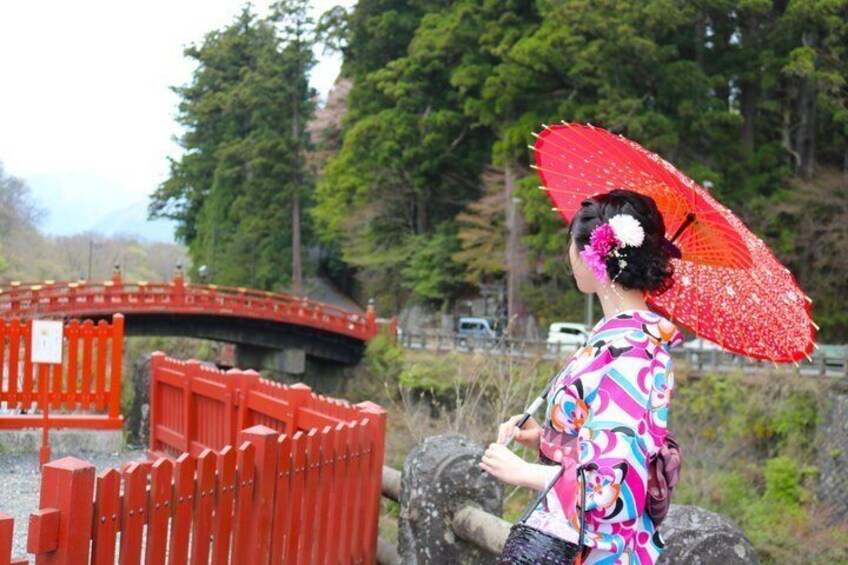 A person in traditional attire with a colorful kimono and a red parasol, standing near a red bridge in a natural setting. 2