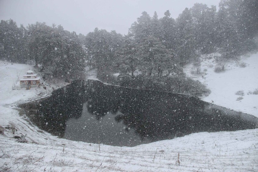 A snow-covered landscape with a pond, trees, and a small building. 4