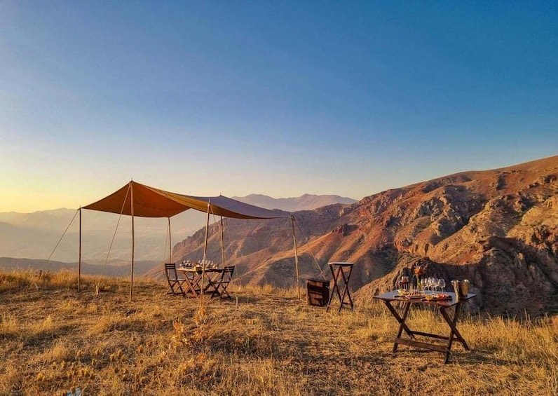 A camping setup with a tent and tables on a grassy hillside with mountains in the background. 1