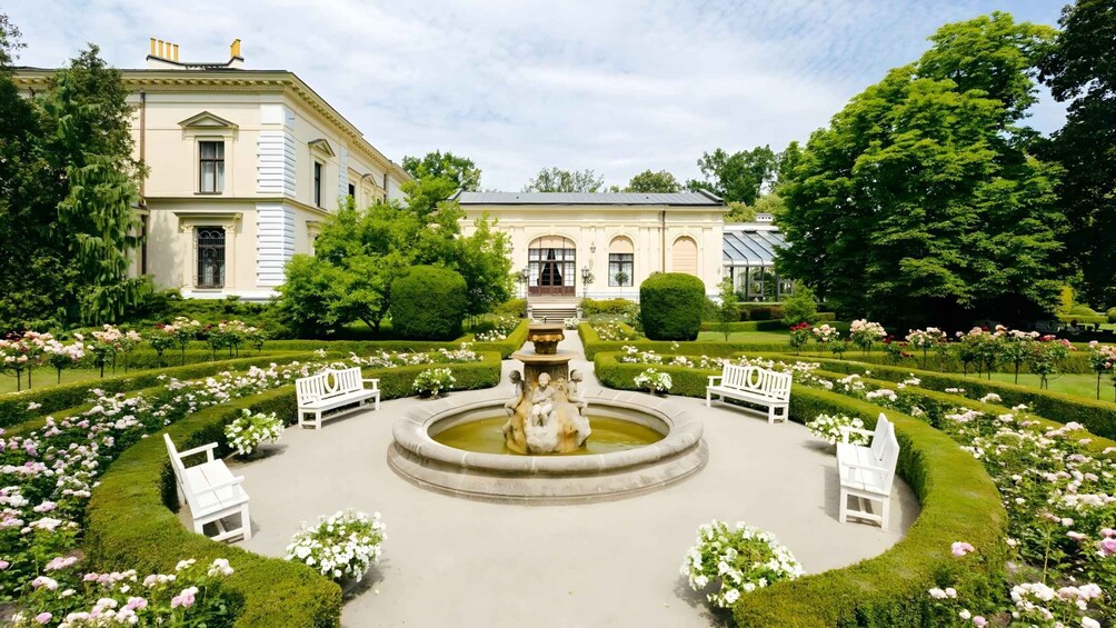 En formel have med en central fontæne, velplejede blomsterbede og klassisk arkitektur. 3