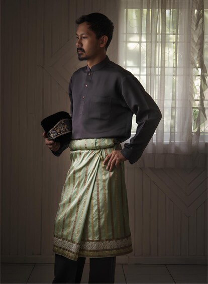 A person in traditional attire standing indoors with a patterned skirt and a dark shirt. 5