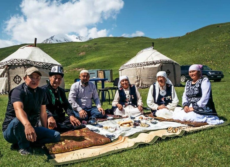 A group of people sitting on a blanket outdoors with yurts in the background. 1