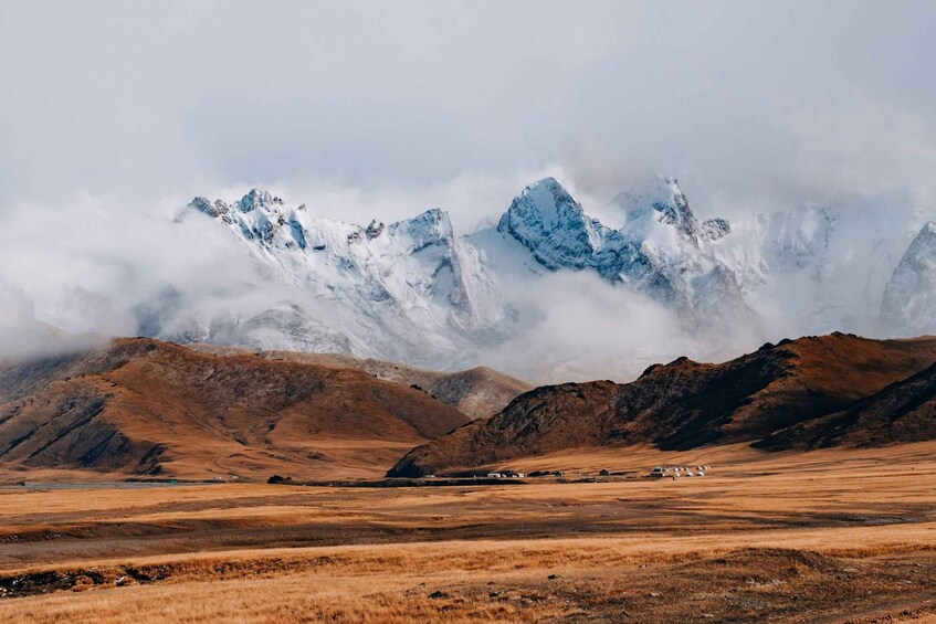 A mountainous landscape with snow-capped peaks, a dry valley, and a few scattered buildings. 3