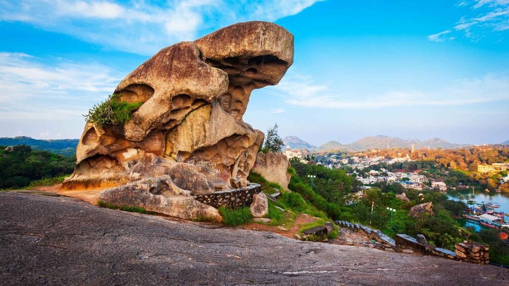 A large rock formation with a unique overhang, a paved path, and a view of a town with buildings and a body of water. 1