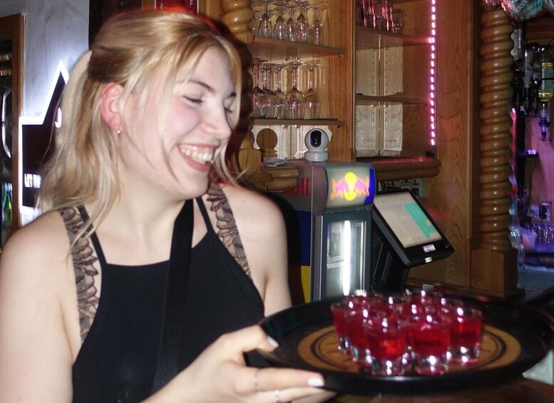 A person holding a tray with red drinks in a bar setting with shelves of glasses and a Red Bull machine. 4