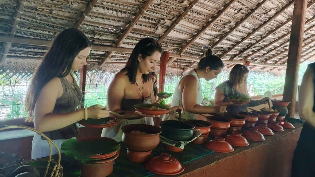 Four women cooking outdoors under a thatched roof, surrounded by clay pots. 2