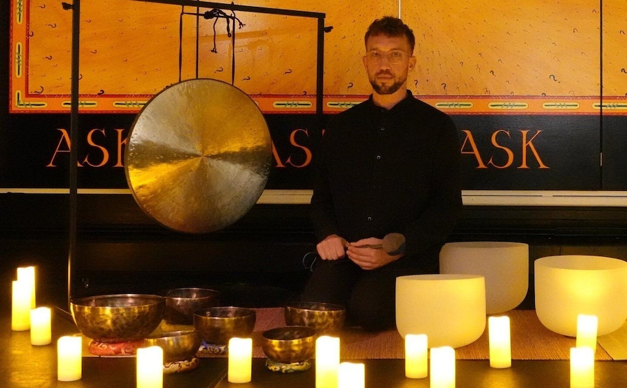 A person seated in front of a room with a large gong, several bowls, and candles. 1