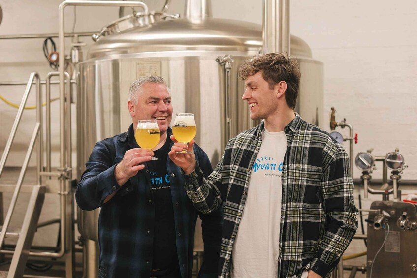Two people holding glasses of beer in a brewery setting with stainless steel tanks and brewing equipment in the background. 1
