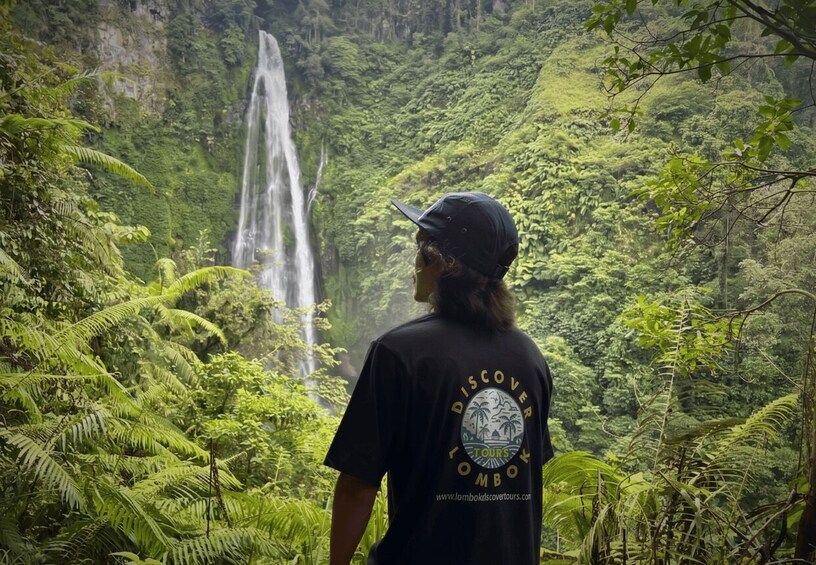 A person wearing a black t-shirt with a logo and text, standing in a lush green forest with a waterfall in the background. 1