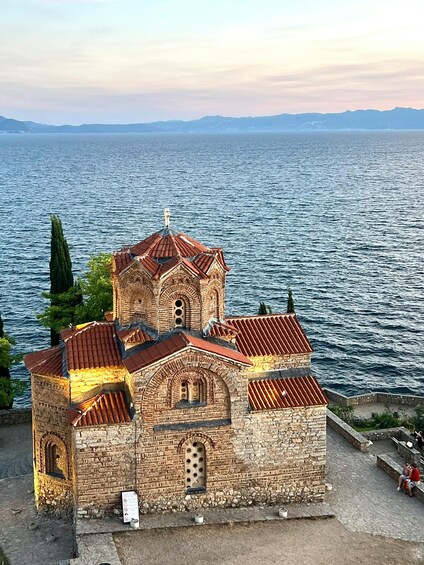 A historic church with a red-tiled roof and stone walls, situated by a large body of water with mountains in the background. 5