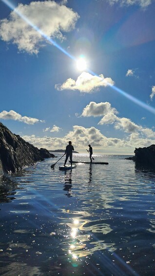 Two people paddleboarding on a calm body of water with rocky cliffs on one side and a clear sky with clouds. 5
