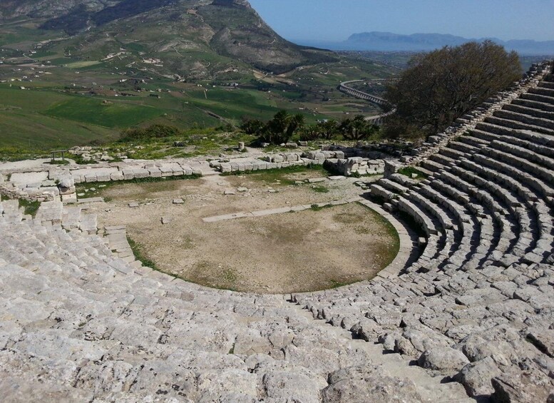 An ancient amphitheater with stone seating and a central stage, surrounded by a hilly landscape. 2