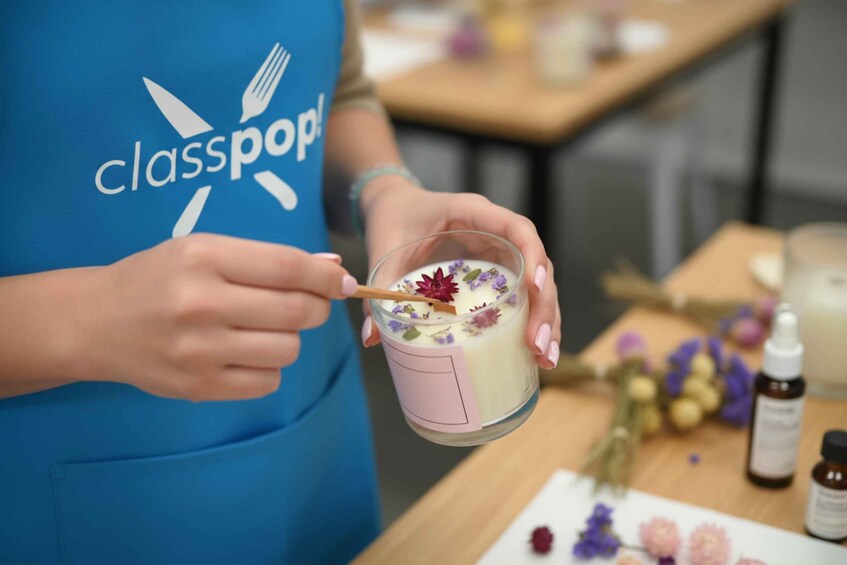 A person in a blue apron with a logo, stirring a drink in a glass with a wooden spoon, with a bouquet of flowers and bottles on a table. 3