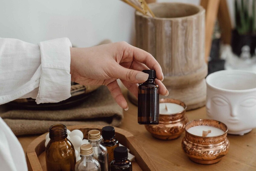A hand holding a small brown bottle, possibly containing essential oils, with other bottles and candles on a wooden surface. 5