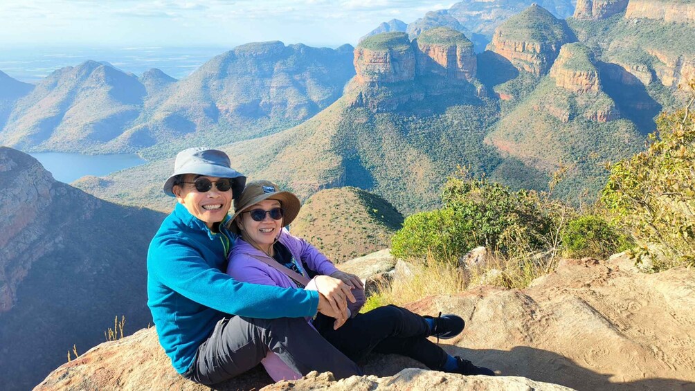 Two people sitting on a rocky outcrop overlooking a mountainous landscape with a valley and a body of water in the distance. 1