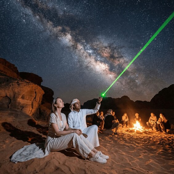A desert night scene with a starry sky, a green laser beam, and people gathered around a campfire. 2