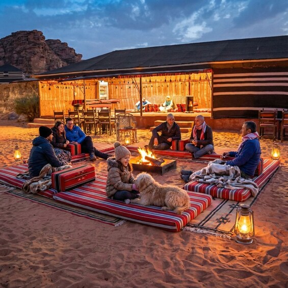 A group of people sitting on striped cushions around a fire pit in a desert setting with a wooden building in the background. 3