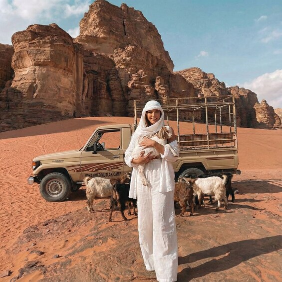 A desert landscape with rocky formations, a beige pickup truck, and a person holding a lamb. 1