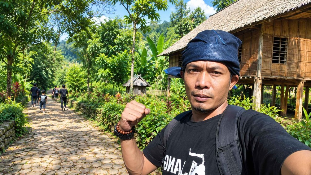 A person taking a selfie with a rural path, a wooden hut, and lush greenery in the background. 3