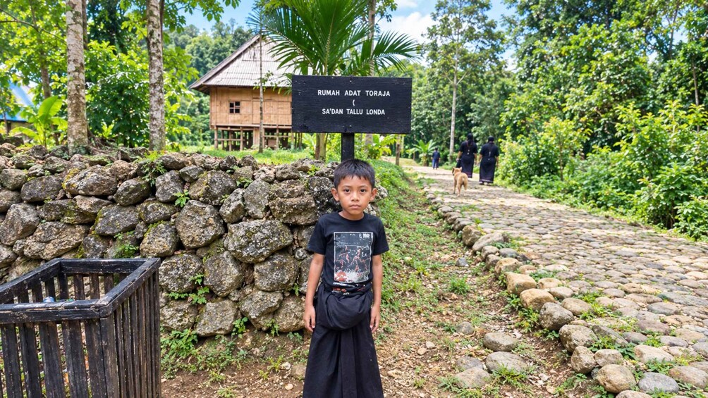 A young boy standing in front of a sign with text, a stone wall, and a pathway leading to a traditional house. 5