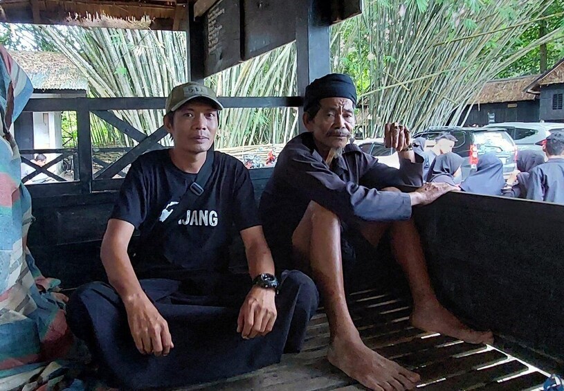 Two men sitting on a bench under a bamboo structure, with a car and a building in the background. 4