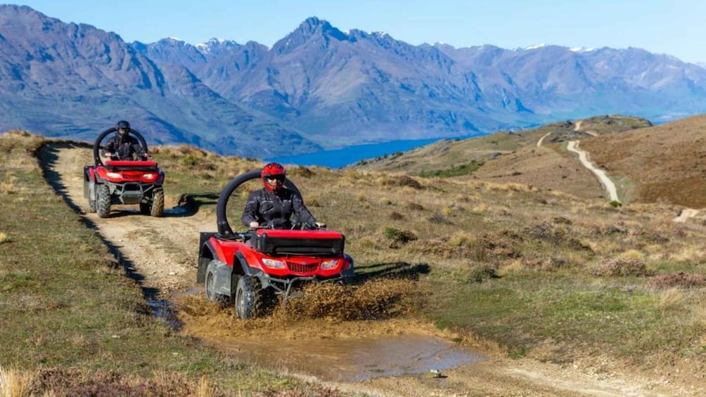 Two people riding red all-terrain vehicles on a dirt path with mountains in the background. 3