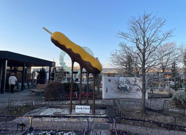 A large yellow and brown ice cream cone sculpture outdoors with a sign reading '#いももフライ' and a tree nearby. 5