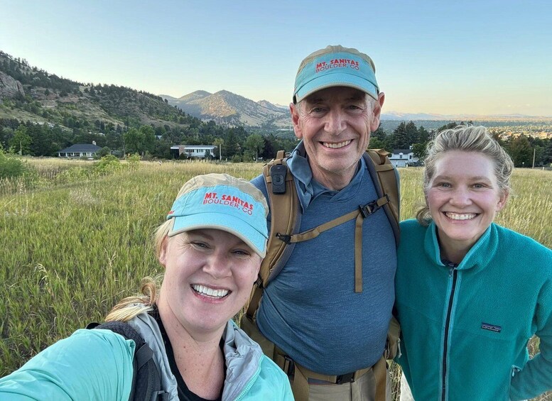 Three people wearing hats with 'MT. SANITAS BOULDER, CO' text, standing in a grassy field with mountains in the background. 5