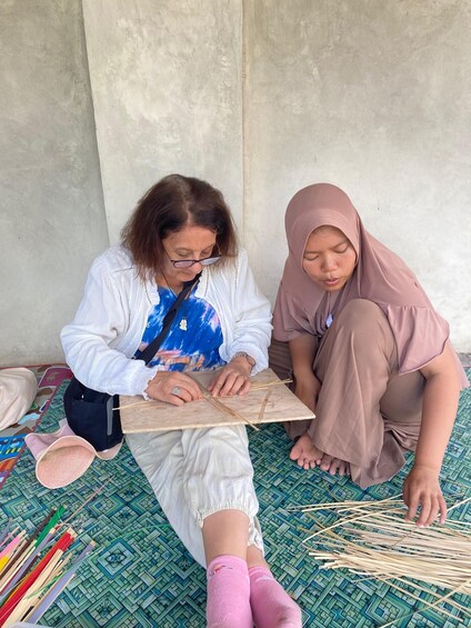 Two individuals seated on a patterned mat, engaged in a craft activity involving wooden sticks and a piece of paper. 3