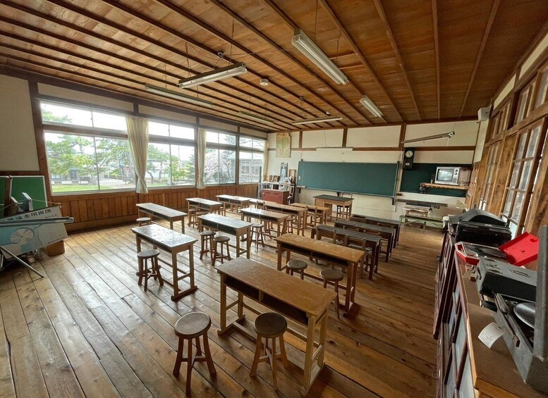 A classroom with wooden desks and chairs, a blackboard, a television, and large windows. 1