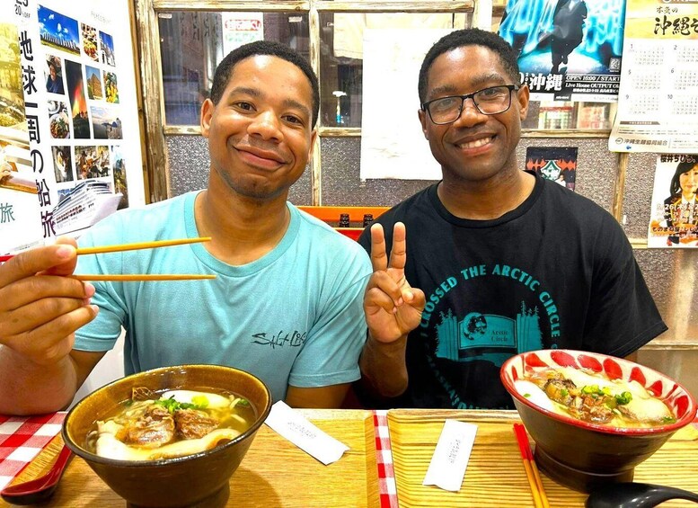 Two people sitting at a table with bowls of soup and chopsticks. 1
