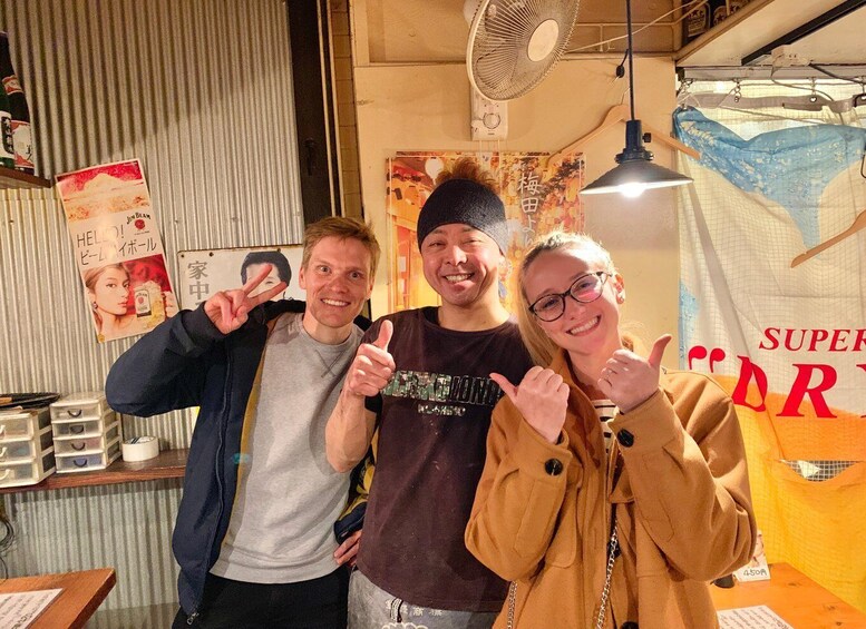 Three people posing in a room with a fan, a poster, and a sign with Japanese text. 3