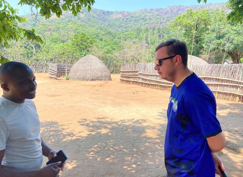 Two people standing outdoors with a traditional thatched structure and a wooden fence in the background. 1