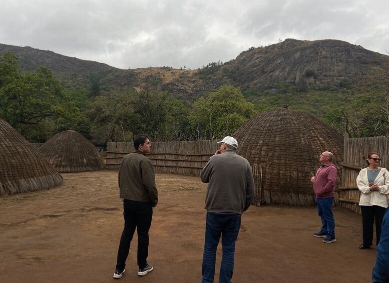 A group of people observing traditional thatched huts in a rural setting with hills in the background. 4