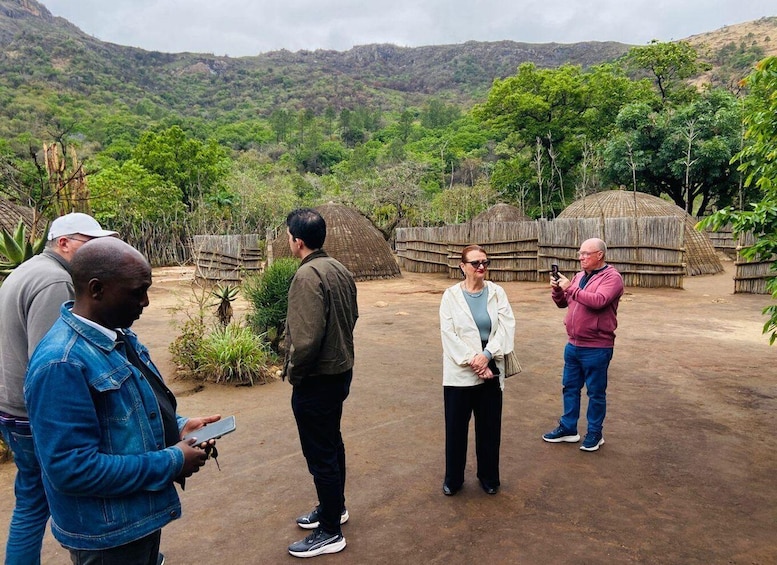 A group of people standing outdoors, some taking photos, with thatched structures and a mountainous landscape in the background. 5