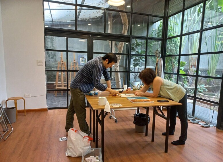 Two people painting at a wooden table in a room with large windows. 3