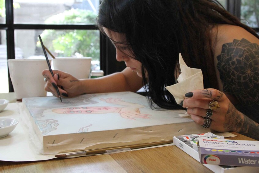 A person painting on a canvas with a paintbrush, seated at a table with art supplies and a cup. 1