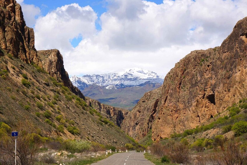 A mountain road flanked by rocky cliffs with a snow-capped peak in the distance. 3